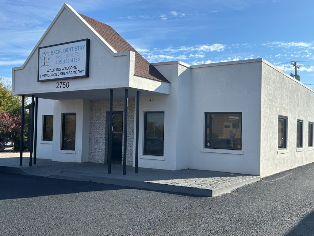 Building with a sign reading 'Excel Dentistry and Braces,' including a phone number, walk-in welcome notice, and same-day emergency service. White exterior with a brown roof and several windows.